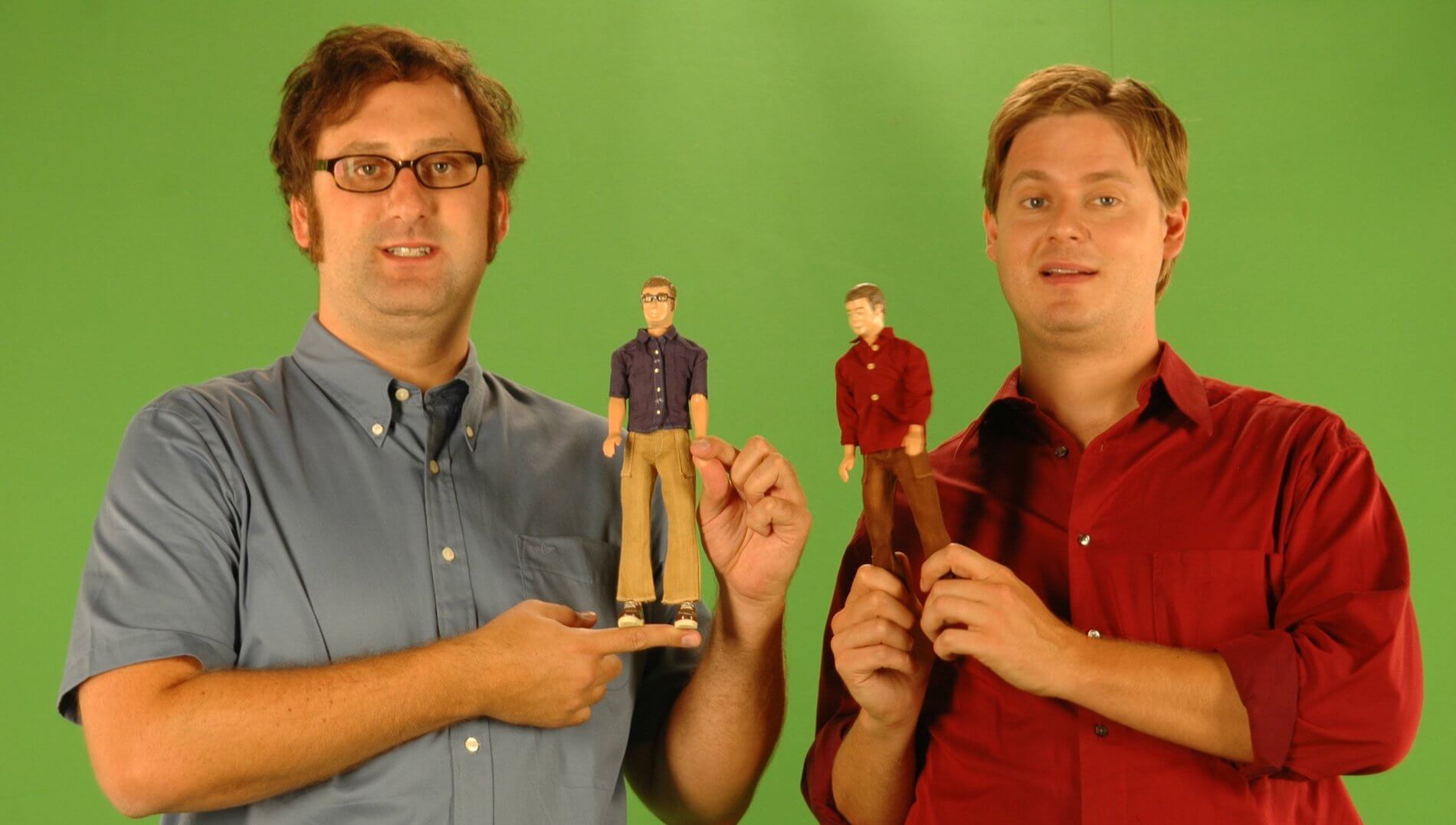 A man with dark hair in a grey dress shirt and another man with blond hair in a red button-up shirt hold doll versions of themselves in a skit for 'Tim and Eric'