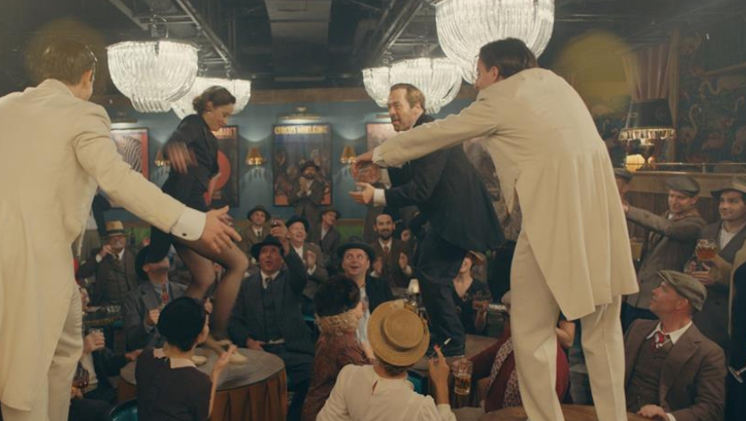 Image of "The Performance" film, with three men and one woman tap dancing on top of tables at a busy venue.